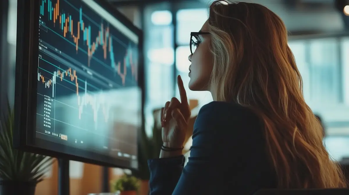 Femme concentrée analysant des graphiques financiers sur un grand écran dans un bureau moderne, avec courbes et indicateurs boursiers visibles.